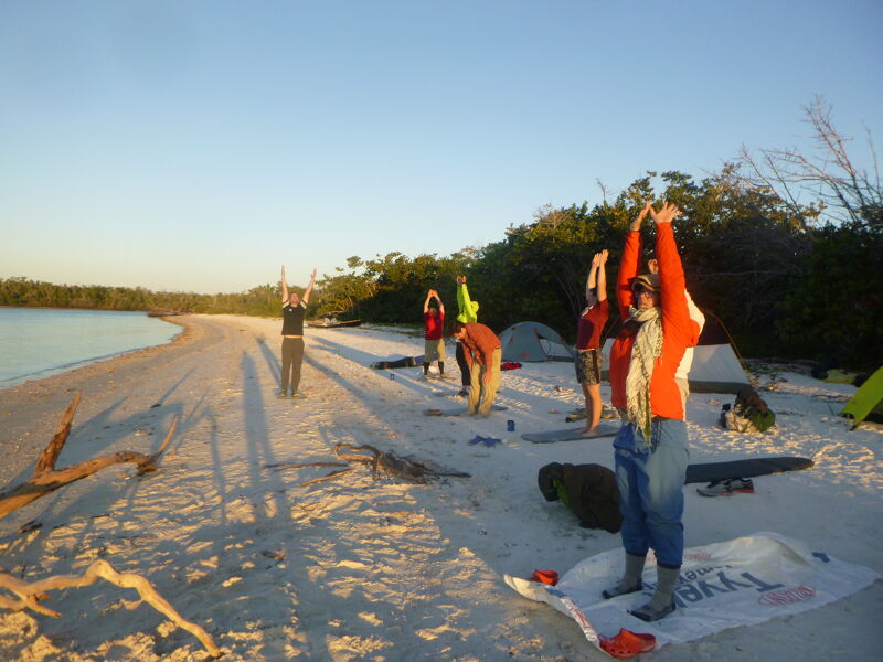 A group of people are doing yoga on a sandy beach near the ocean. Some tents are pitched nearby, suggesting they might be camping. The sun is shining, casting long shadows on the sand. The overall scene suggests a peaceful and active morning by the sea.